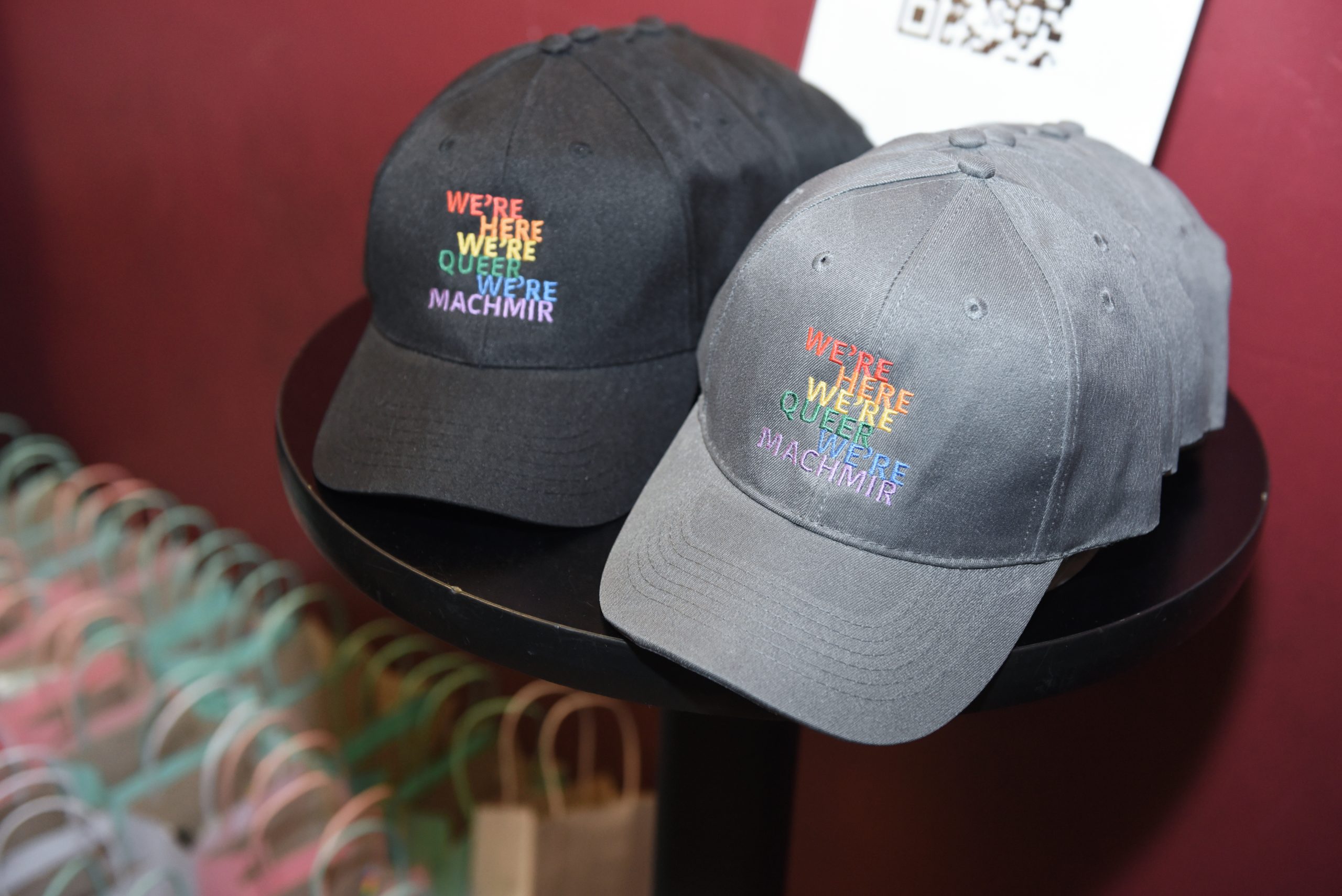 Black and Gray Baseball caps with "We're Here, We're Queer, We're Machmir" embroidered on them in rainbow letters